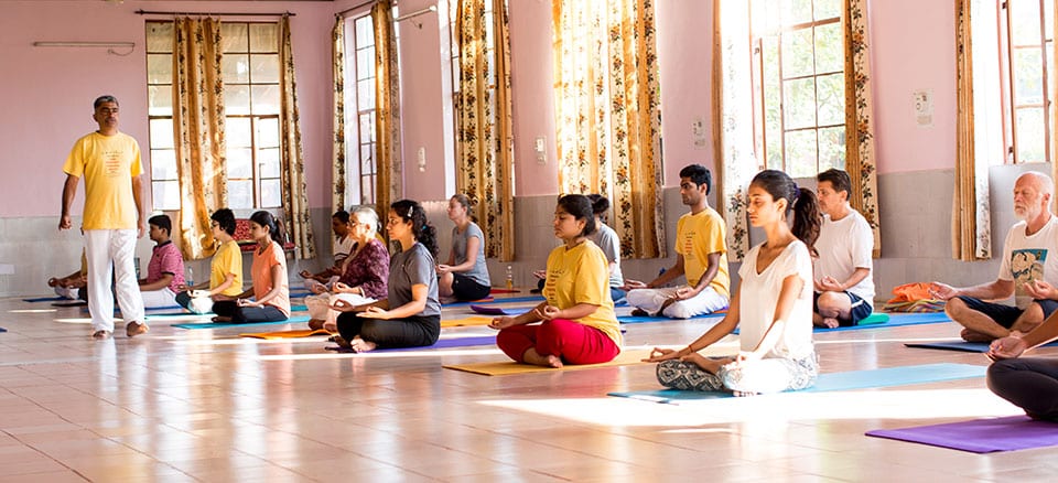 a group yoga class seated in meditation on mats in a bright hall.