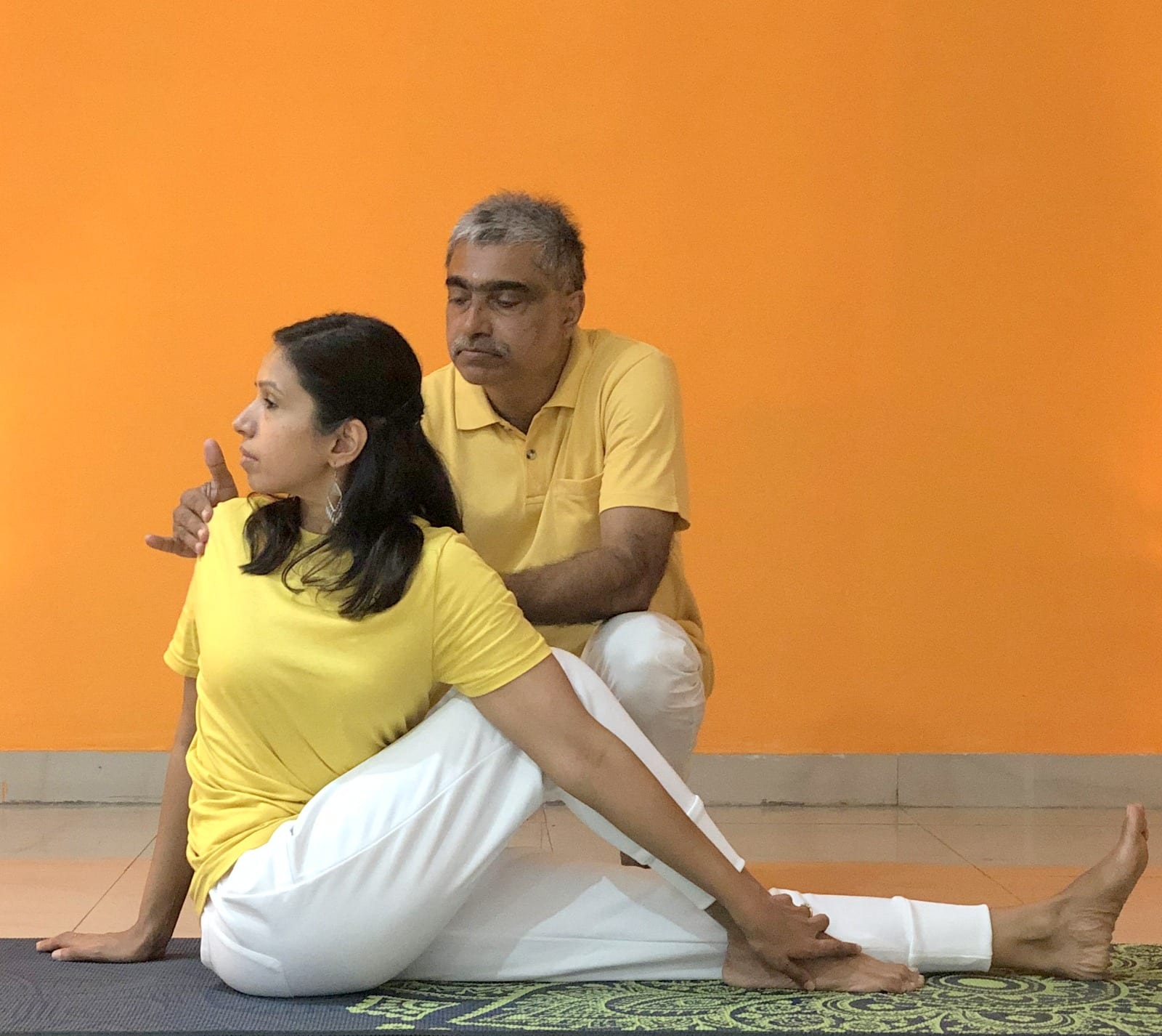 A teacher kneeling behind a seated student, guiding her deeper into Ardha Matsyendrasana, the Half Spinal Twist, on a mat in front of an orange wall