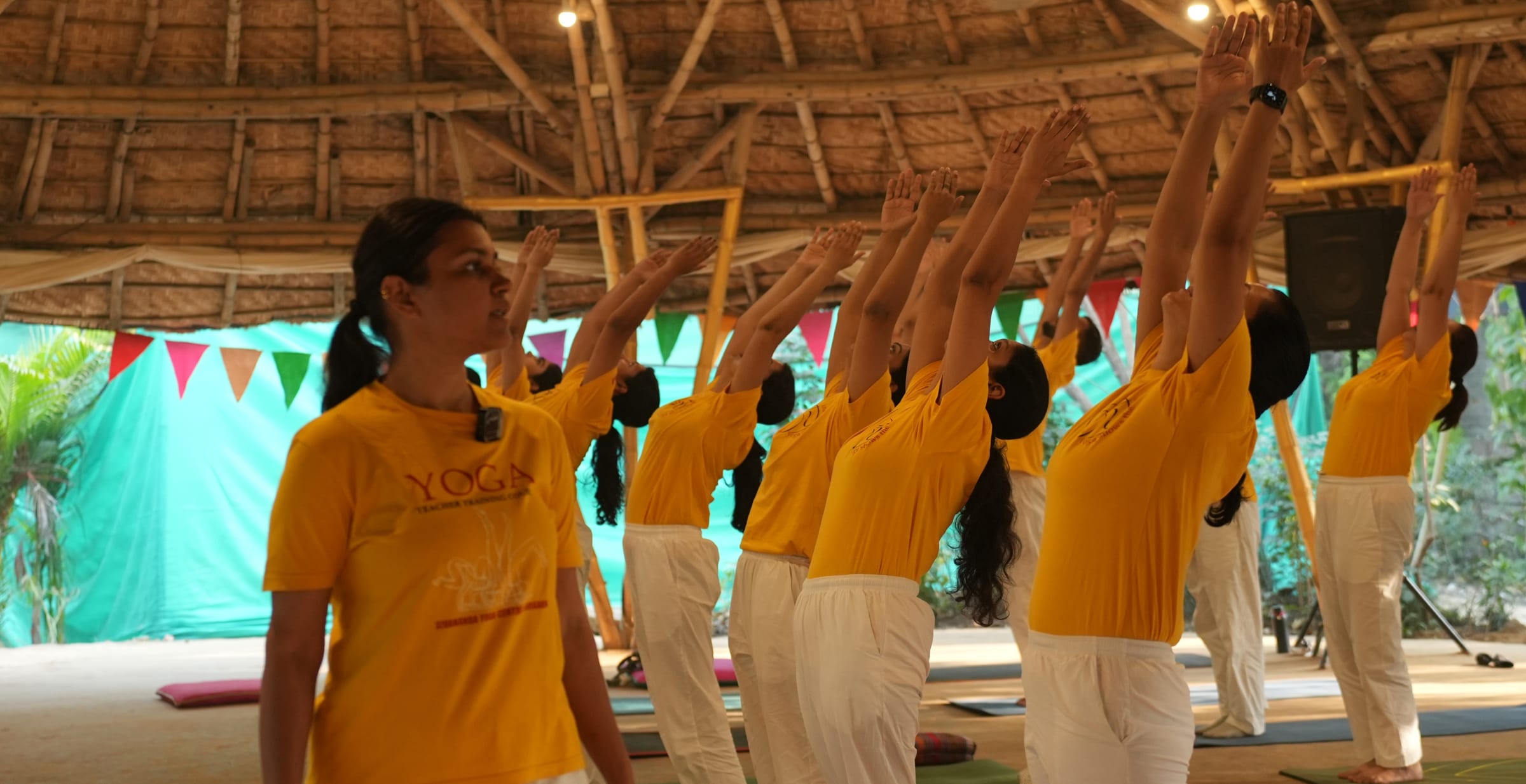 A class standing with both arms overhead under the thatched pavilion roof at a Sivananda Gurgaon retreat