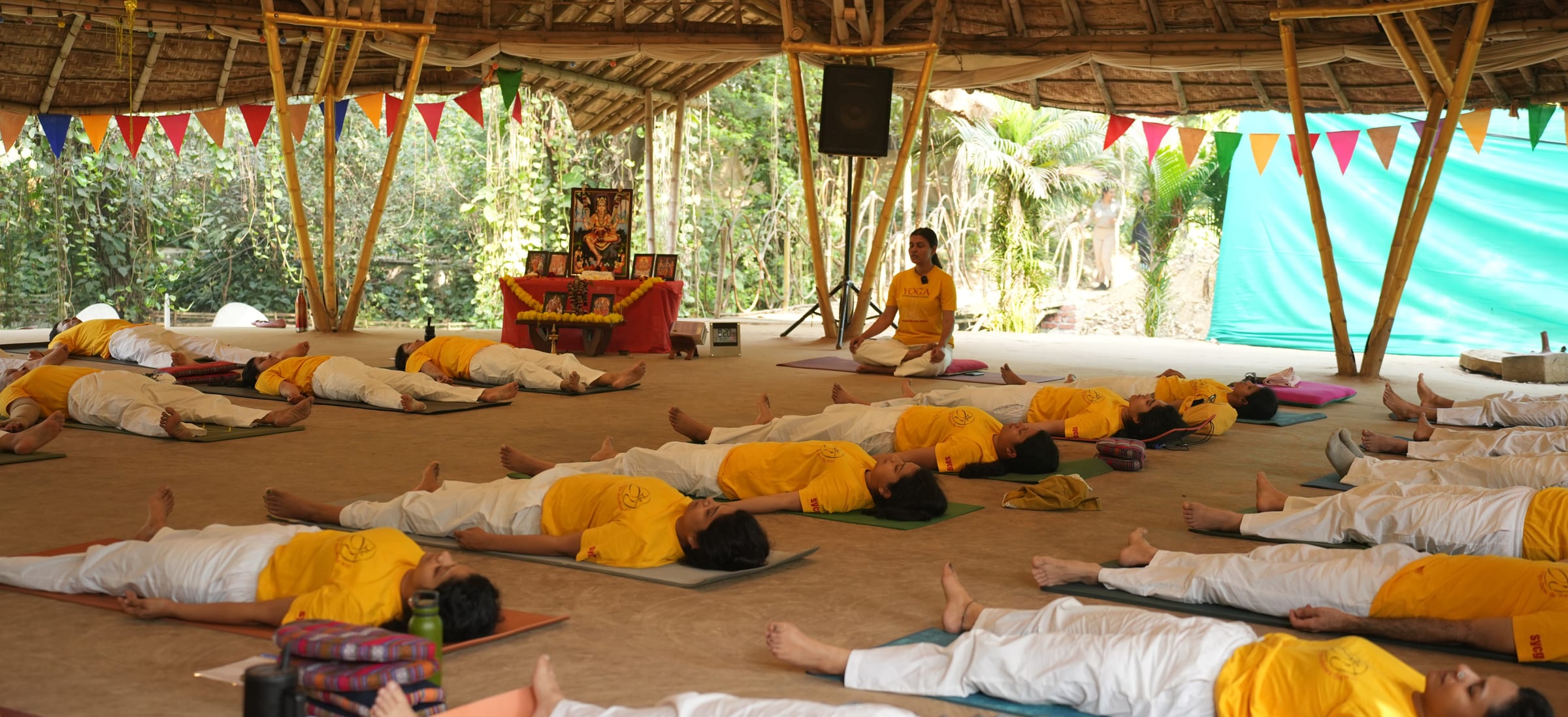 A full class lying in Savasana on mats under the pavilion, a teacher seated before the altar at the far end