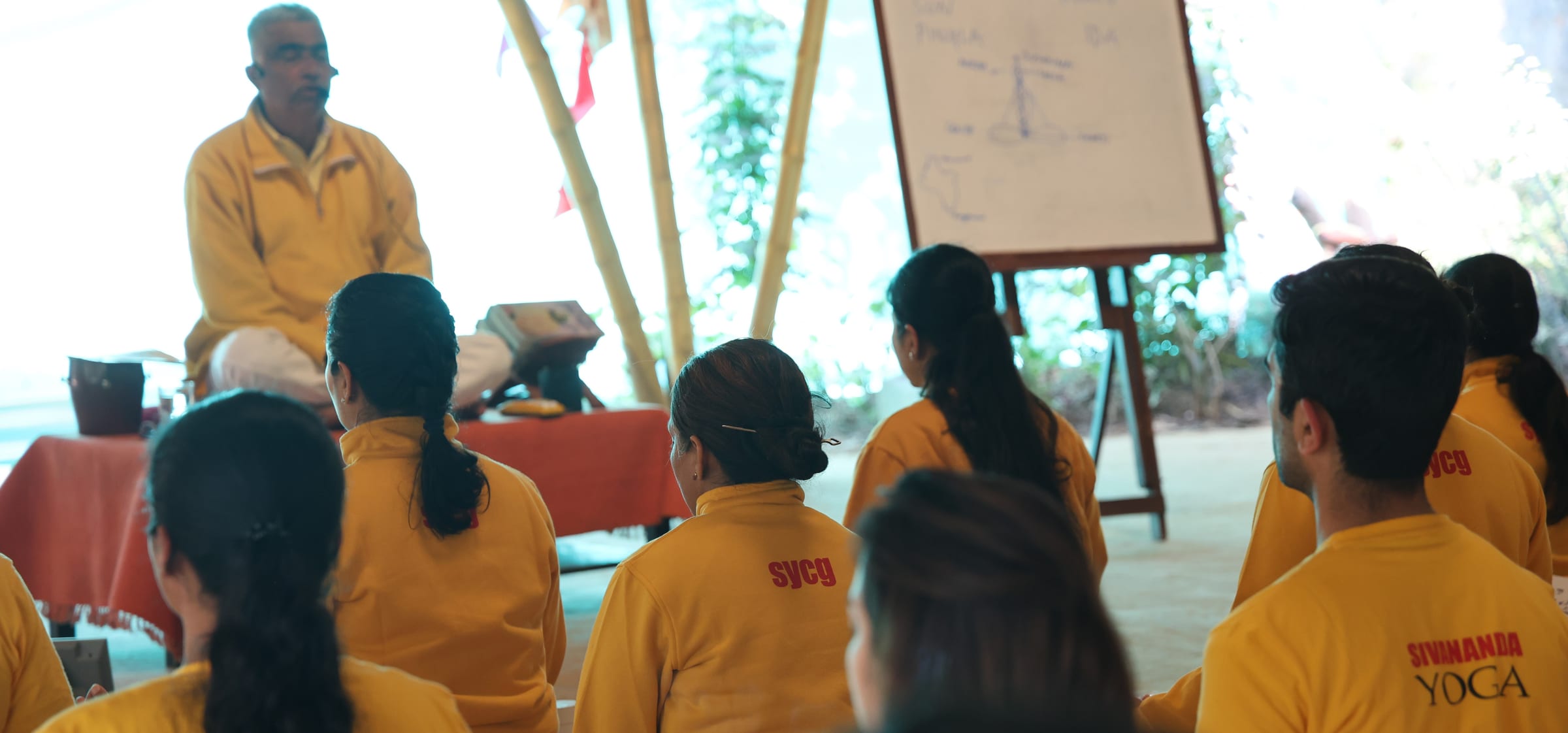 Arun Pandala speaking from a raised seat to a group seated on the floor, a flip chart beside him