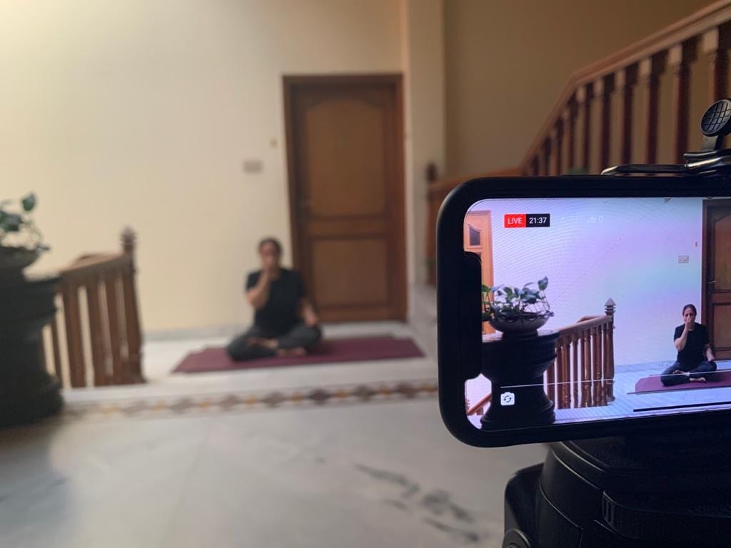 A phone on a tripod filming a live class, the teacher seated on her mat at the foot of a staircase and framed on its screen.