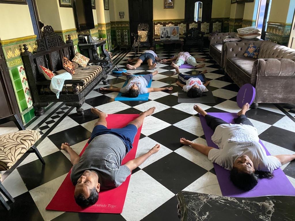Two students in Savasana on mats on a chequered floor at home.