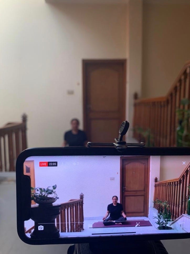 A phone on a tripod carrying a live class, the teacher seated on her mat in the hall behind it.
