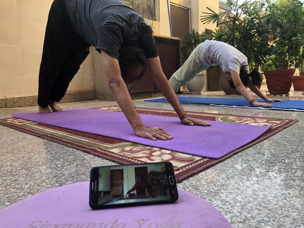 Three students in Downward Dog on mats at home, a phone on the front mat carrying the class.