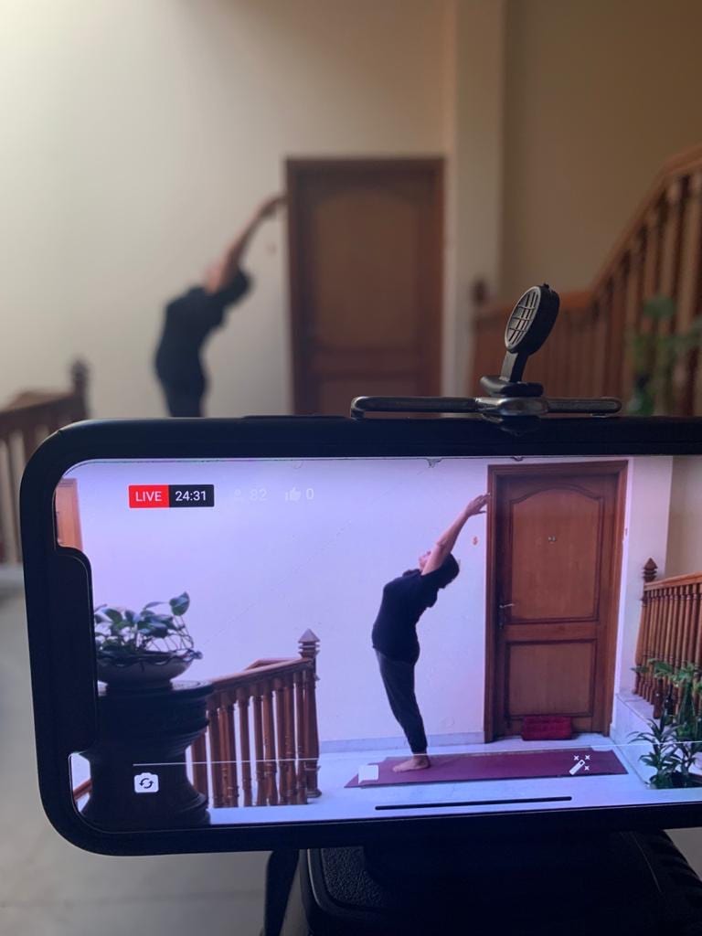 A phone on a tripod carrying a live class, the teacher standing in a side bend and framed on its screen.
